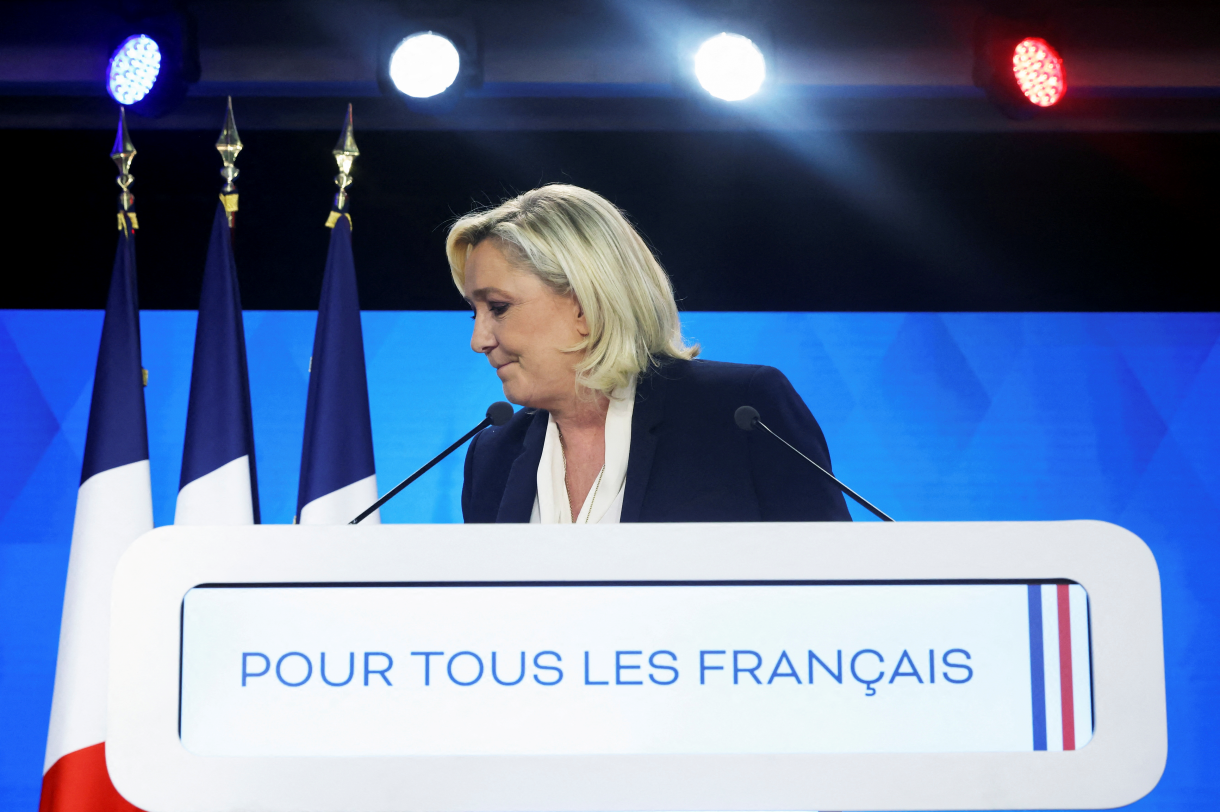 Marine Le Pen in front of a podium looking to her right with French flags. 