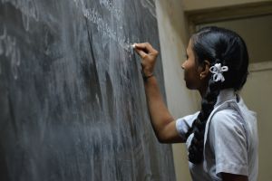 A young Indian girl writes on a chalkboard in a classroom