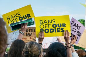 Hands hold yellow sign saying "Bans off our bodies" in front of a blue sky