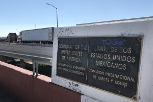 A sign marking the US-Mexico border, with semi trucks driving in the background