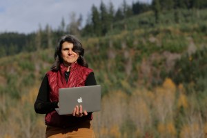 A woman holding a laptop stands outside observing her farm.