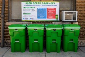 Four green scrap drop off bins sit in front of a brick wall