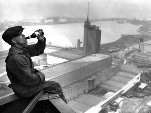 An iron worker sits on the edge of a skyscraper while drinking a beverage