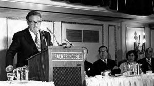 A black and white photo of Henry Kissinger speaking to a large crowd.