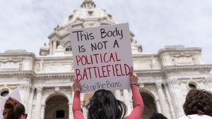 A sign reading "This body is not a political battlefield" is held before a capitol building and cloudy sky.