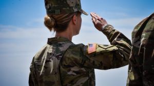 A woman in uniform faces away from camera and salutes in front of a blue sky.