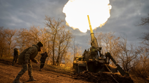 a cannon fires with flames and soldiers stand in the foreground, bare trees in the background in Ukraine.