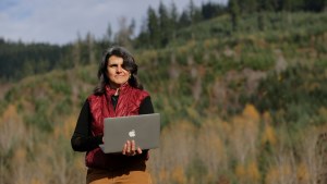 A woman holding a laptop stands outside observing her farm.
