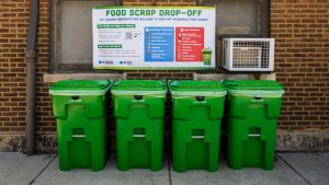 Four green scrap drop off bins sit in front of a brick wall